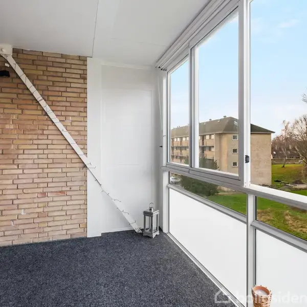 Altan med tæppebelægning er omgivet af glasvægge, hvor udsigten viser en græsplæne og træer. En lanterne står i hjørnet, mens murstensvæg og en skråstiver er synlige.