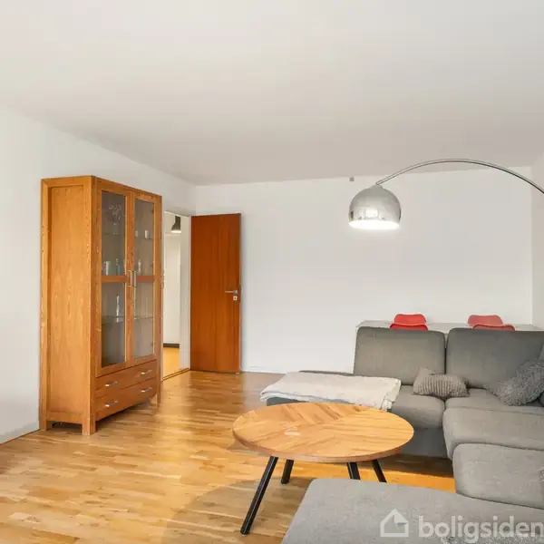 Gråt sofaarrangement med en buet lampe over, placeret i en stue med trægulv. TV står på en skænk til venstre, og et vitrineskab står ved en væg.