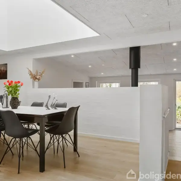 Spisestue med træbord og sorte stole står på trægulv. Bordet er udsmykket med blomster. Baggrunden viser en moderne, lys indendørs ramme med store vinduer, der fører udenfor.