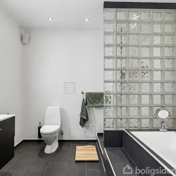 Badeværelse med glasbyggesten væg ved badekar, hvidt toilet, mørke skabe under håndvask, grå fliser på gulvet, grønne håndklæder på væggen, i et moderne, minimalistisk design.