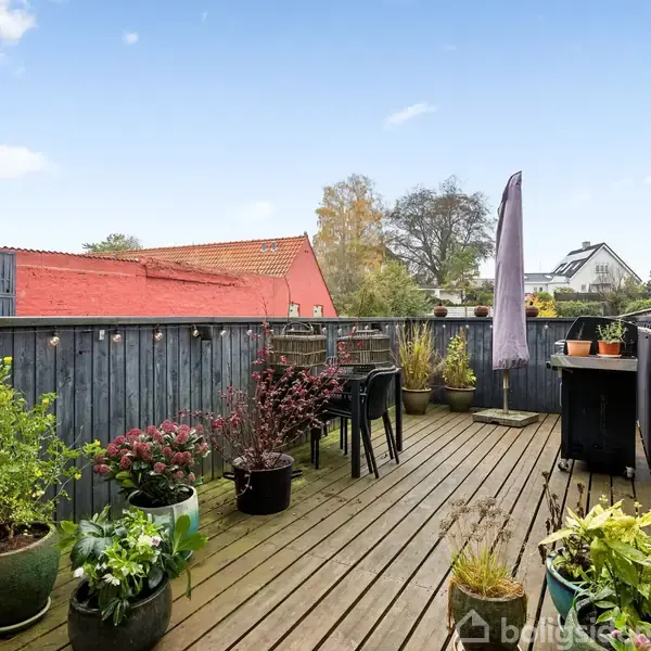 En træterrasse er fyldt med forskellige planter i krukker. Et bord med stole står midt på terrassen. En åben udsigt viser træer og hustage i baggrunden.