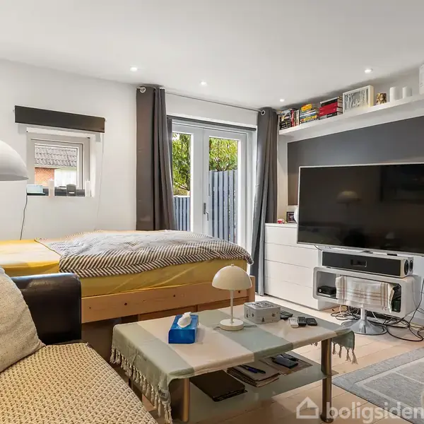 Stue med sofa og sofabord, der har en lampe og bøger. En seng står ved et vindue med udsigt til en gårdhave, og et TV hænger på en sort væg.
