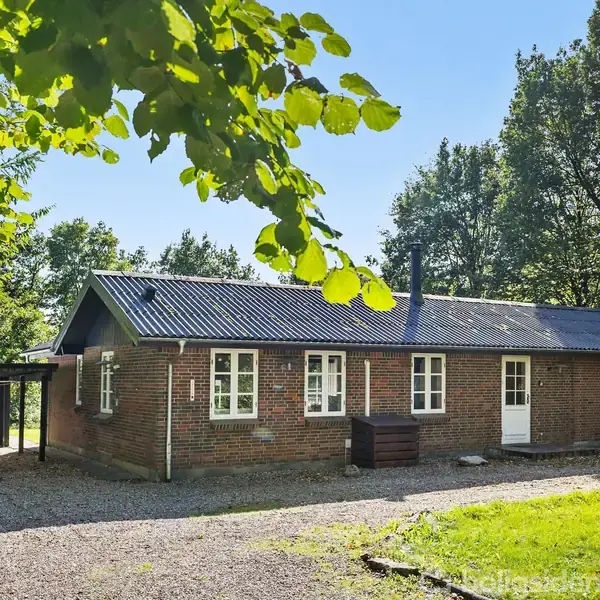 Et murstenshus med sort tag står stille omgivet af træer og grønt græs. Gruset indkørsel leder til en carport ved siden af.