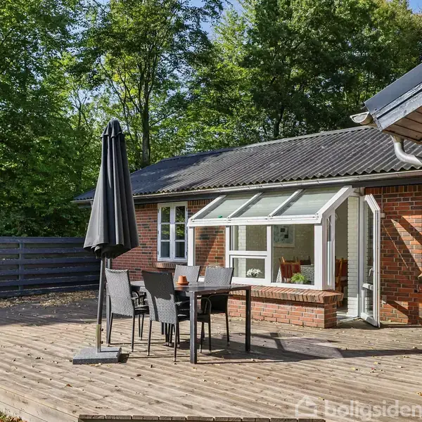 En åben terrasse med et bord og sorte stole under en stor parasol. Terrassen tilhører et rødstenshus med store vinduer, omgivet af frodig skov.