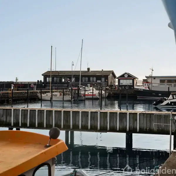 Både ligger ved kajen i en havn. Forklaringsskilte på bygninger ses. To både er fortøjet ved en træbro. Vandet er roligt, og en bygning ses i baggrunden.