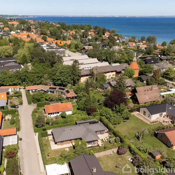Luftfoto af en kystnær landsby viser huse med røde tage og grønne haver langs en vej. Baggrunden består af træbevoksning og en blå havudsigt.
