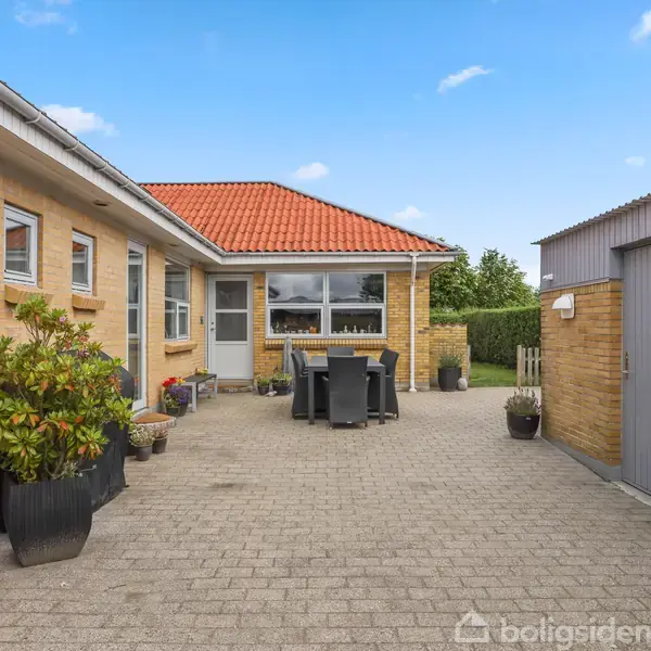 Rødt tegltag over gul murstenshus set fra en gårdhave med flisebelægning, havemøbler og plantekrukker. Tilstødende bygning med gråt træskur. Klar blå himmel i baggrunden.