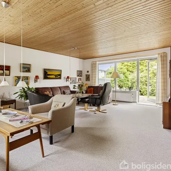 Stue med brune og beige møbler, herunder en sofa og lænestole. Malerier og planter dekorerer rummets vægge. Store vinduer og en glasdør fører til en terrasse med udsigt til grønne træer.