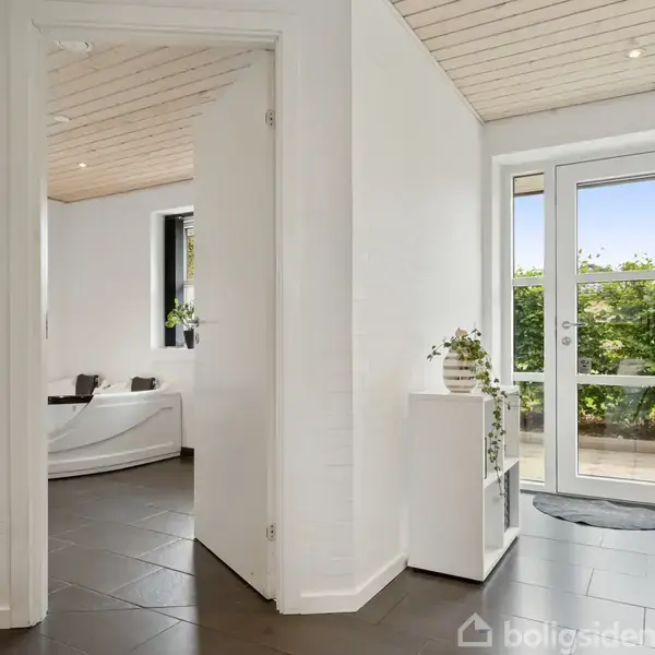 Dør står åben mod terrasse, trin fører ud til fliser med grøn beplantning udenfor. Indendørs er rummeligt med hvide vægge og loft, sort flisegulv og badeværelse på venstre side.