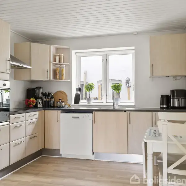Grå sovesofa står i en stue med træbund, dekoreret med en pelset tæppe og puder. En stribet vase med blomster står på et glasbord, omgivet af lette vægge og to vinduer.