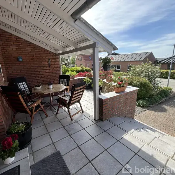 Et sæt havesæt står ubenyttet på en overdækket terrasse med udsigt til en blomstrende have og en rolig vej.