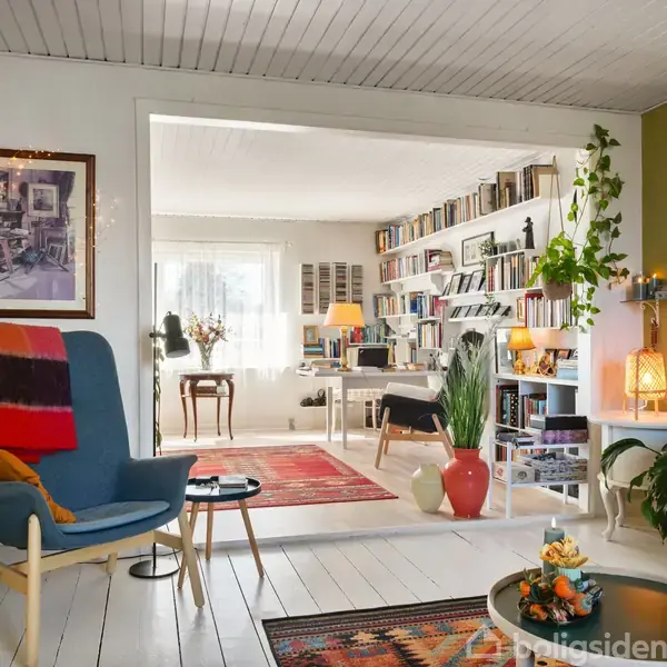En stue med en blå lænestol med farverigt tæppe, står ved siden af et lille bord. Rummet åbner sig til et arbejdsområde med boghylder og en skrivebordslampe, omgivet af planter og tæpper.