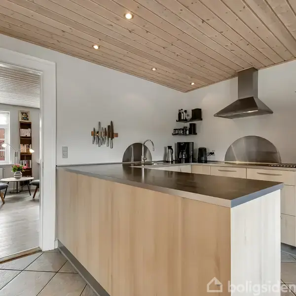 Køkkenet har en stor køkkenø og moderne apparater, herunder en ovn og emhætte. Det ligger ved siden af en lys stue med møbler og store vinduer.