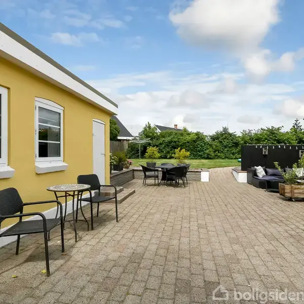 En gul bygning står stille ved en flisebelagt terrasse med borde og stole. Ved siden af er en grøn have omkranset af buske og træer under en blå himmel.