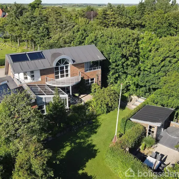 En stor moderne villa med solpaneler på taget står blandt tæt bevoksede træer. En græsplæne og et lille anneks ses i baghaven, omgivet af frodig natur.