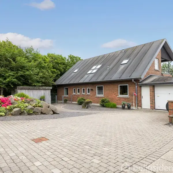 Et rødt murstenshus med en skråtag, liggende i en have med brolagt indkørsel og blomsterbede, omgivet af træer og buske. Der er en tilknyttet garage.