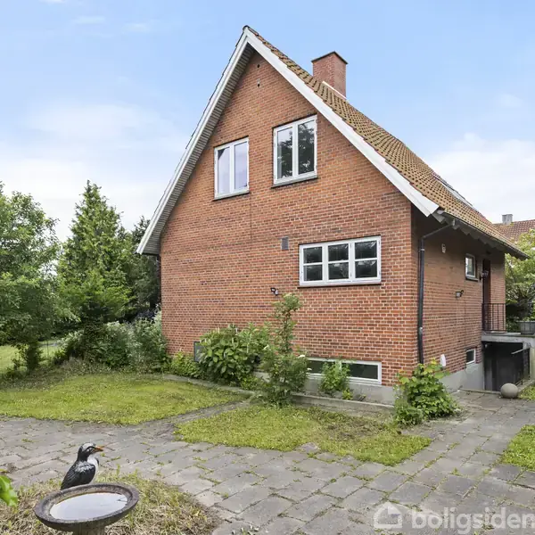Et rødt murstenshus står omkranset af en frodig have. Huset har skråtag og flere vinduer. I forgrunden ses en stenbelagt sti og en fuglefigur ved et fuglebad.