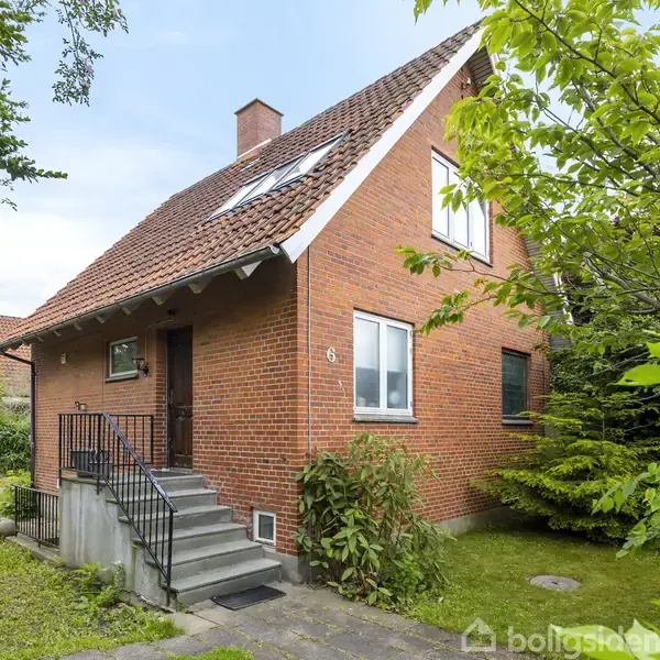 Rødt murstenshus står stille omgivet af frodig have. Trappe fører til hoveddør under skråtag. Adresse på facade: "6". Græsplæne og træer i baggrunden.