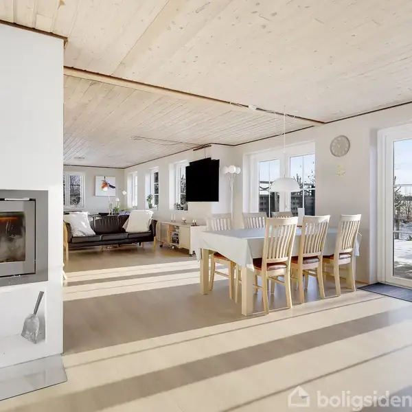 Brændeovn opvarmer stue med spisebord og stole, mens solen skinner gennem store vinduer. Rummet har lyse trægulve og loft, og udsigt til sneklædt landskab udenfor.