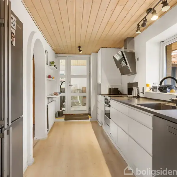 Køkken med hvide skabe og stål-køleskab. Køkkenet er ryddeligt, med moderne apparater og vindue mod gårdhave. Loftet er af lyst træ, og belysning inkluderer spotlights.