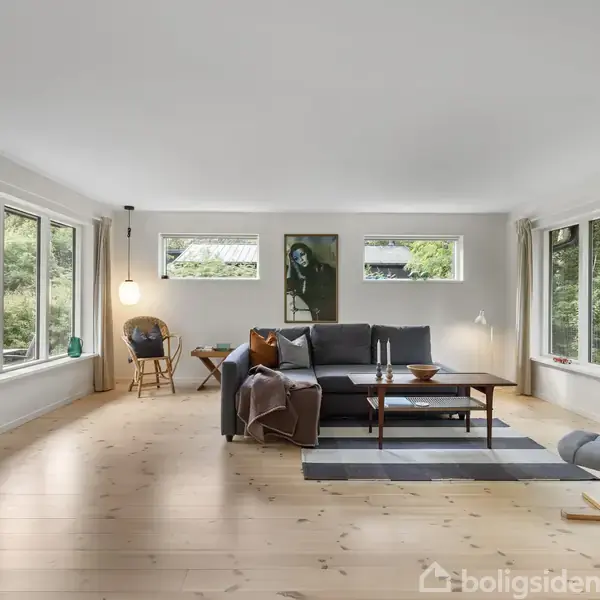 En stue med en sort sofa og bord på et tæppe. Store vinduer omgiver rummet, hvilket giver udsyn til grønne omgivelser. En lænestol står i den højre del af rummet.