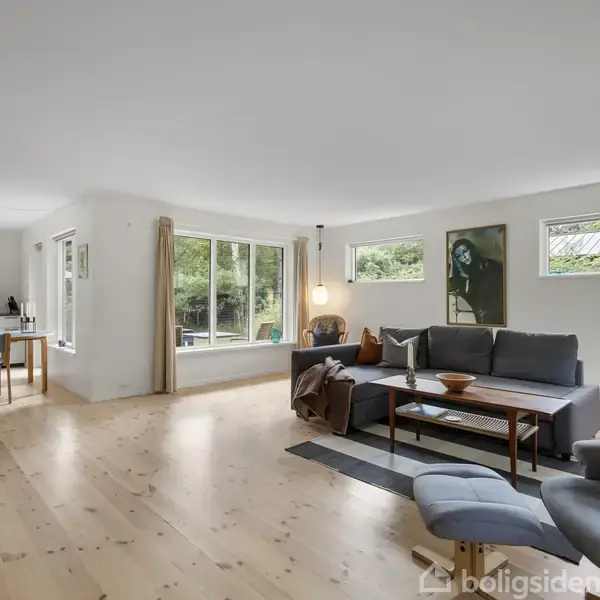 En moderne stue med grå sofa og lænestol, placeret i et lyst rum med lyse trægulve. Til venstre ses et spisebord med stole nær store vinduer, der slipper naturligt lys ind.