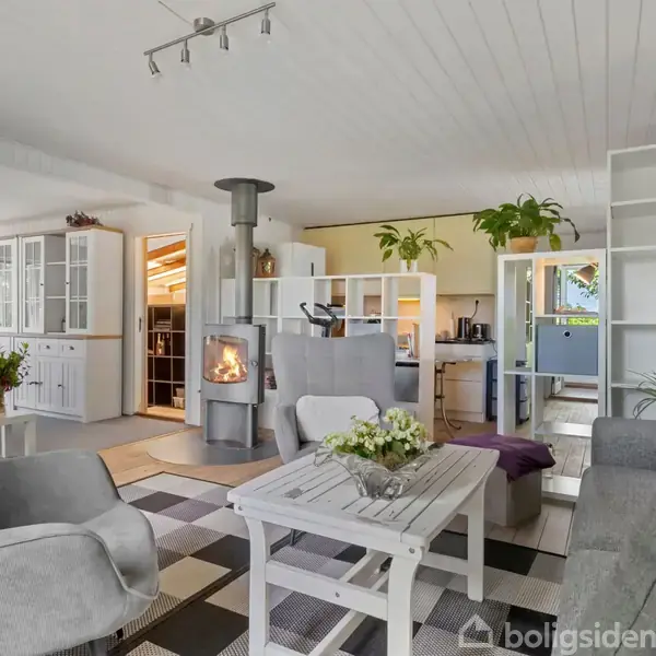 En hyggelig stue med en brændeovn i funktion, omgivet af komfortable møbler. Rummet har et lyst, moderne design med gulv-til-loft hylder og planter, hvilket giver en varm atmosfære.