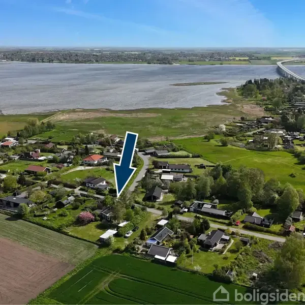 En blå pil peger på et hus i en landlig by omgivet af marker og skov, tæt ved en bred flod med en lang bro i baggrunden.