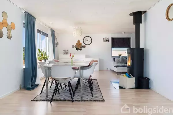 Spisebord med hvide stole står på et tæppe i en lys stue med træovn. Blå gardiner, planter og vægdekorationer skaber en hyggelig atmosfære. Væggen har en ur.