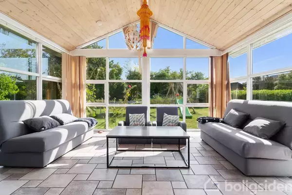 To grå sofaer står overfor hinanden med et sort sofabord imellem sig. Store vinduer giver udsigt til en have med grønne træer og blå himmel udenfor.