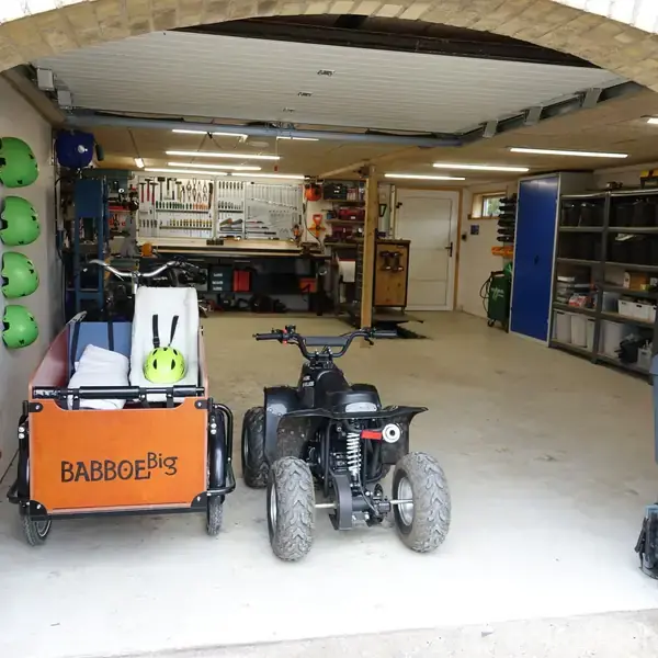 En garage rummer en ladcykel mærket "BABBOE Big" og en ATV; i baggrunden ses værktøj på væggen, hylder med opbevaring og grønne hjelme hængende.