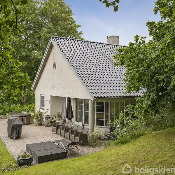 Et hvidt hus med gråt tegltag står i en grøn have. Ved siden af huset er der en terrasse med havemøbler og grilludstyr.