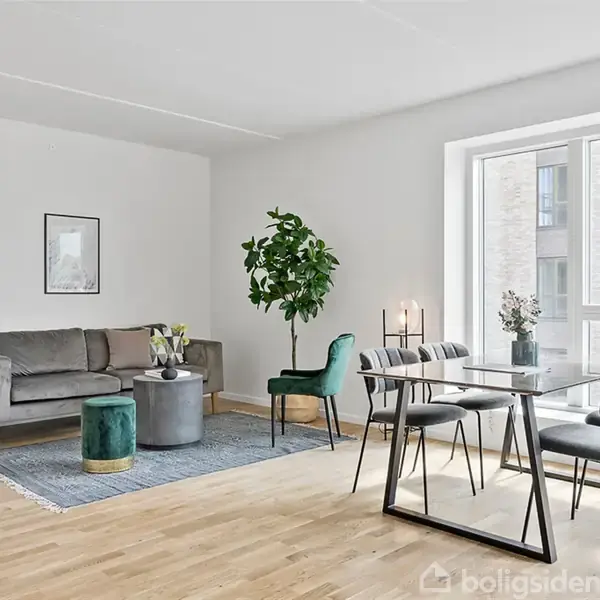 En lys stue med en grå sofa og grøn plante. Et spisebord med stole står ved et stort vindue. Enkle dekorationer skaber en moderne atmosfære.