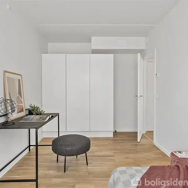 Et værelse med en hvid garderobe mod væggen, et skrivebord med en globe og planter, og en rund skammel på trægulv. En seng med tæppe er i forgrunden.