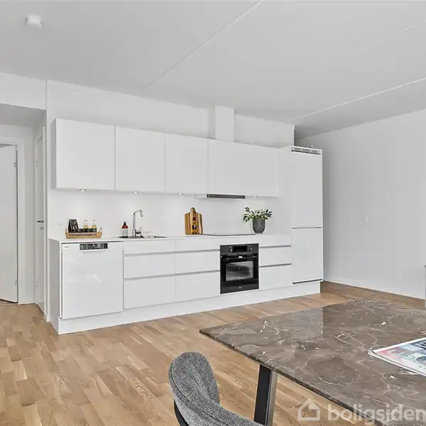 Køkken i moderne stil med hvide skabe integreret i en åben stue. Spisebord med tidsskrift og stol foran. Trægulve og dekorative planter kompletterer rummet.