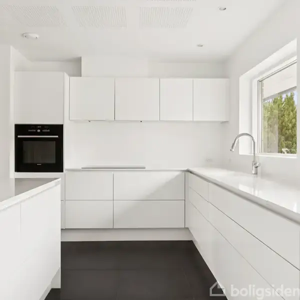 Et moderne køkken med hvide skabe og integreret ovn. En stor vask med armatur er ved vinduet. Rummet oplyses af naturligt lys.