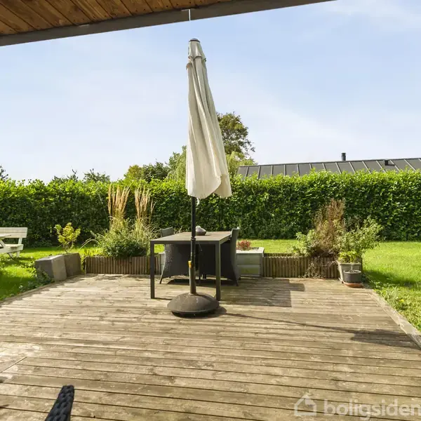 Terrasse med træbrædder har et bord med en sammenklappet parasol. Omkring er der planter, en græsplæne og et træskur. Hækken skaber en grøn baggrund under en klar himmel.