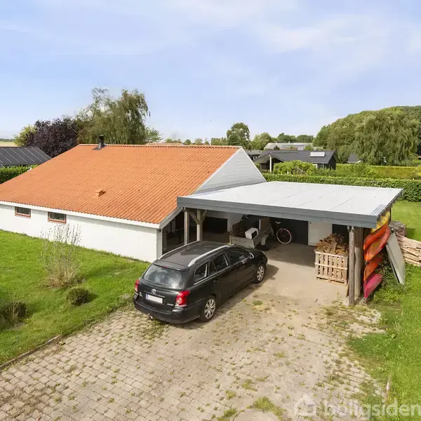Et hus med orange tag står ved siden af en carport, hvor en sort bil er parkeret. Flere kajakker er opbevaret under et halvtag. Græsplæne og træer omgiver området.