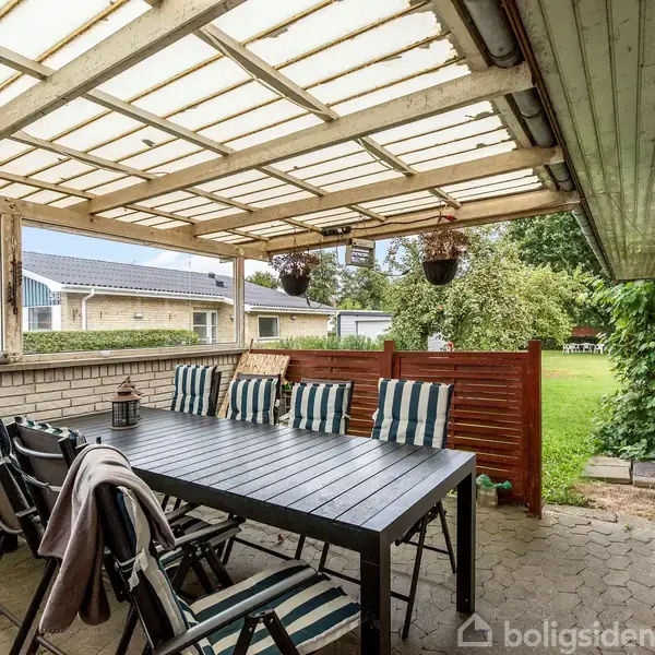 Et stort, mørkt havebord med stribede stole står under en overdækket terrasse. På bordet er en lanterne, og i baggrunden ses en grøn have omgivet af huse.