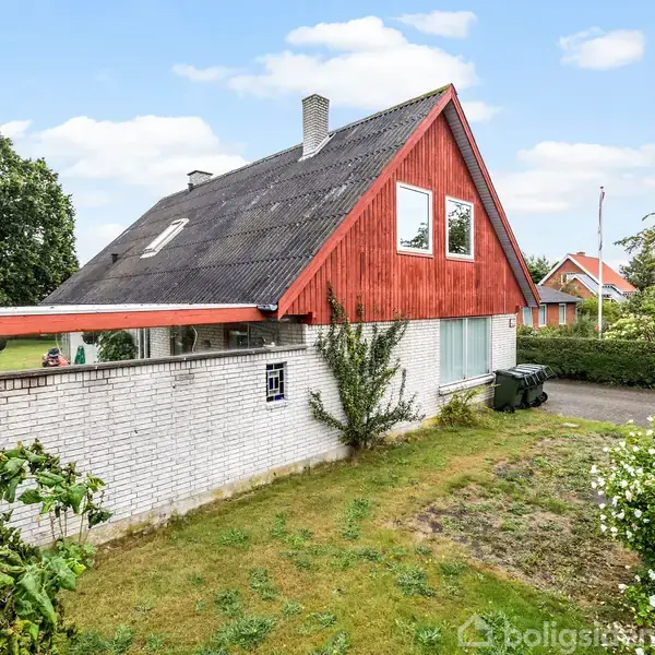 Et rødt træhus med sort tag står ved siden af en hvid mur i en grøn have. Huset er omgivet af buske og træer under en blå himmel med skyer.