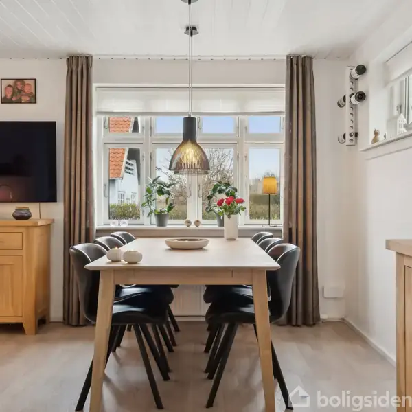 Et spisebord i træ med seks sorte stole står i et lyst rum. En lampe hænger over bordet, omgivet af et stort vindue med gardiner. På væggen er et fjernsyn og billeder, og der er kommoder på begge sider.