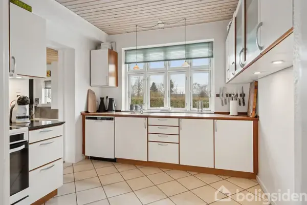 Køkken med hvide skabe og sort komfur. Kaffemaskine står på bordet. Flisegulv og brunt trægulv ses. Lys gang fører til dør med udsigt til haven.