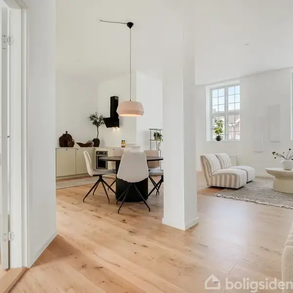 Lyst stue- og køkkenområde med trægulv. Et bord med stole står nær et åbent køkken. Store vinduer lader lys ind i rummet med lyse møbler og planter.