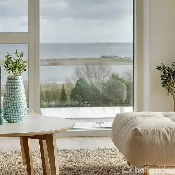 Et rundt bord står i et lyst rum med en vase med planter og sorte lysestager, ved siden af en beige ottoman. Store vinduer viser udsigt over vand og natur.