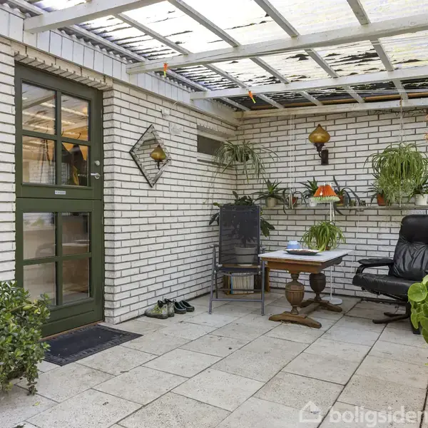 En overdækket terrasse med hvidkalkede murstensvægge, hvor en sort lænestol står ved et rundt træbord. Grønne planter hænger og står i potter omkring i rummet.