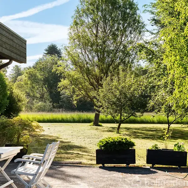 Terrasse med hvide havemøbler står ved siden af et grønt græsareal med træer. Skoven ses i baggrunden under en blå himmel med spredte skyer.