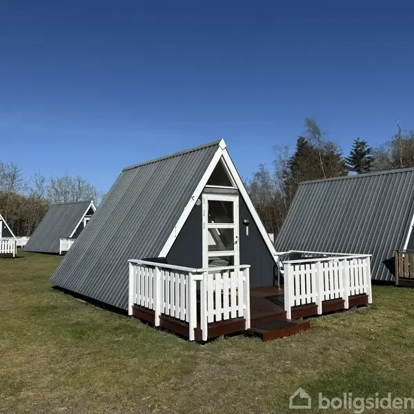 Tre A-formede træhytter står stille på en græsmark. Hytterne har hvide døre og træhegn omkring små verandaer. Baggrunden viser træer mod en blå himmel.