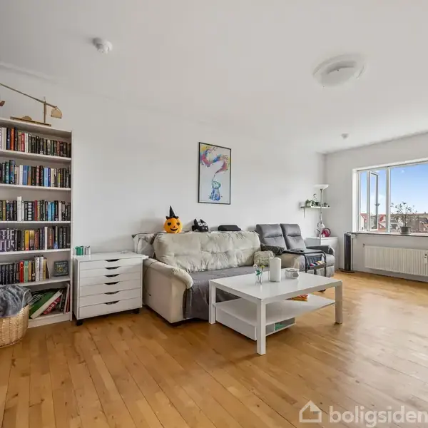 En stue med en beige sofa og et sofabord i midten. Ved siden af sofaen er en fyldt bogreol, og i baggrunden ses et stort vindue, som giver lys.