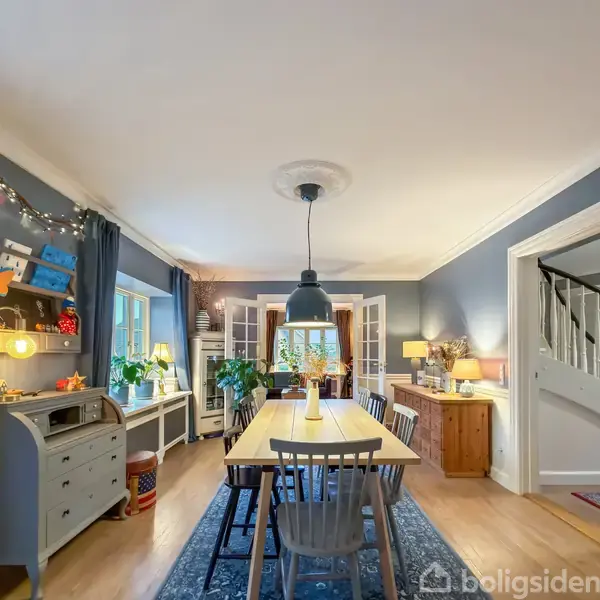 Spisebord med stole står centralt i et lyst spisestue, omgivet af lamper, reoler med pynt og planter, samt hvide døre i baggrunden, der fører til en anden stue.