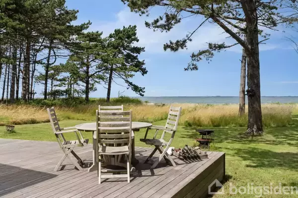 Træmøbler står på en terrasse; stole omringer et rundt bord. I baggrunden er der græsplæne, træer og havudsigt under en lys blå himmel.